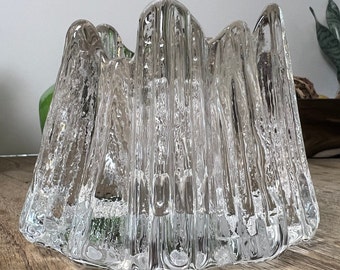 May include: A clear glass candle holder with a textured, rippled surface. The holder has a unique, organic shape with multiple peaks and valleys.