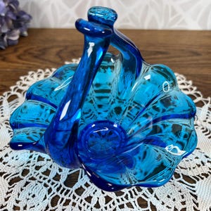 May include: A blue glass candy dish with a handle in the shape of a basket. The dish has a scalloped edge and a textured surface.