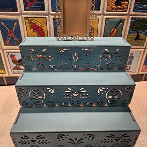 May include: A three-tiered teal display stand featuring intricate laser-cut patterns including skulls, hearts, and floral motifs along the front edges. A small, rectangular, gold-colored ornate frame with a filigree border sits on the top tier.