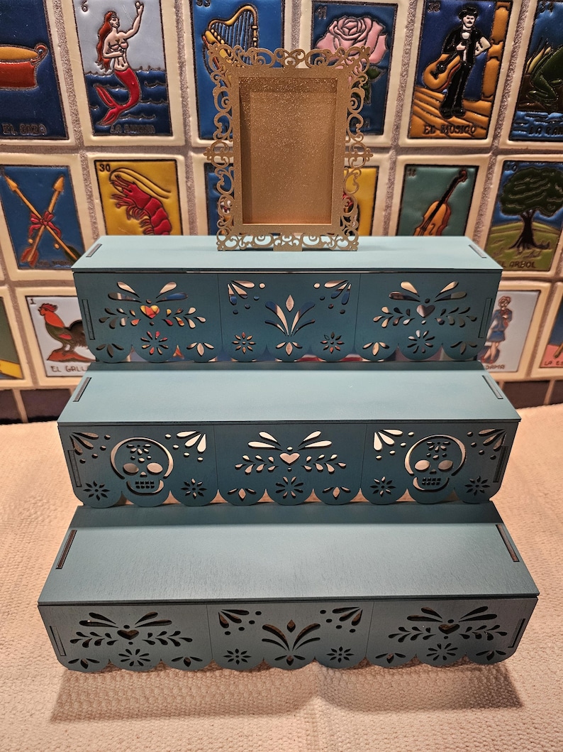 May include: A three-tiered teal display stand featuring intricate laser-cut patterns including skulls, hearts, and floral motifs along the front edges. A small, rectangular, gold-colored ornate frame with a filigree border sits on the top tier.