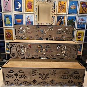 May include: A three-tiered wooden display stand featuring laser-cut decorative edges with floral patterns and two skull motifs on the middle tier. A rectangular frame with an ornate, filigree-style border is attached to the top tier.