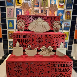 May include: A three-tiered wooden display stand decorated with traditional Mexican Day of the Dead imagery. The stand is painted red and features intricate cutouts of flowers, skulls, and the phrase "Dia de los Muertos".