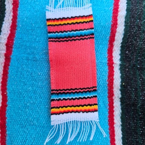 May include: A small, pink woven rug with white, black, yellow, and blue stripes. The rug has fringe on both ends.