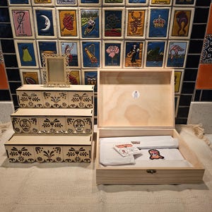 May include: A three-tiered light wood display stand with laser-cut floral and skull motifs, accompanied by an open wooden box containing white tissue paper, a small rectangular card, and a colorful skull sticker with the text 'La Catrina'.