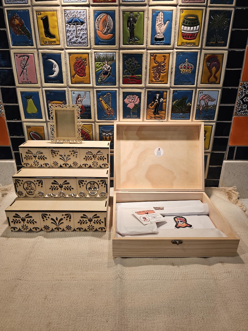 May include: A three-tiered light wood display stand with laser-cut floral and skull motifs, accompanied by an open wooden box containing white tissue paper, a small rectangular card, and a colorful skull sticker with the text 'La Catrina'.