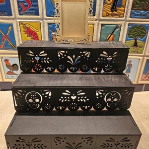May include: A three-tiered black wooden display stand featuring intricate laser-cut patterns along the front edges, including skull motifs on the middle tier. A small, rectangular, gold-colored ornate frame sits centered on the top tier.