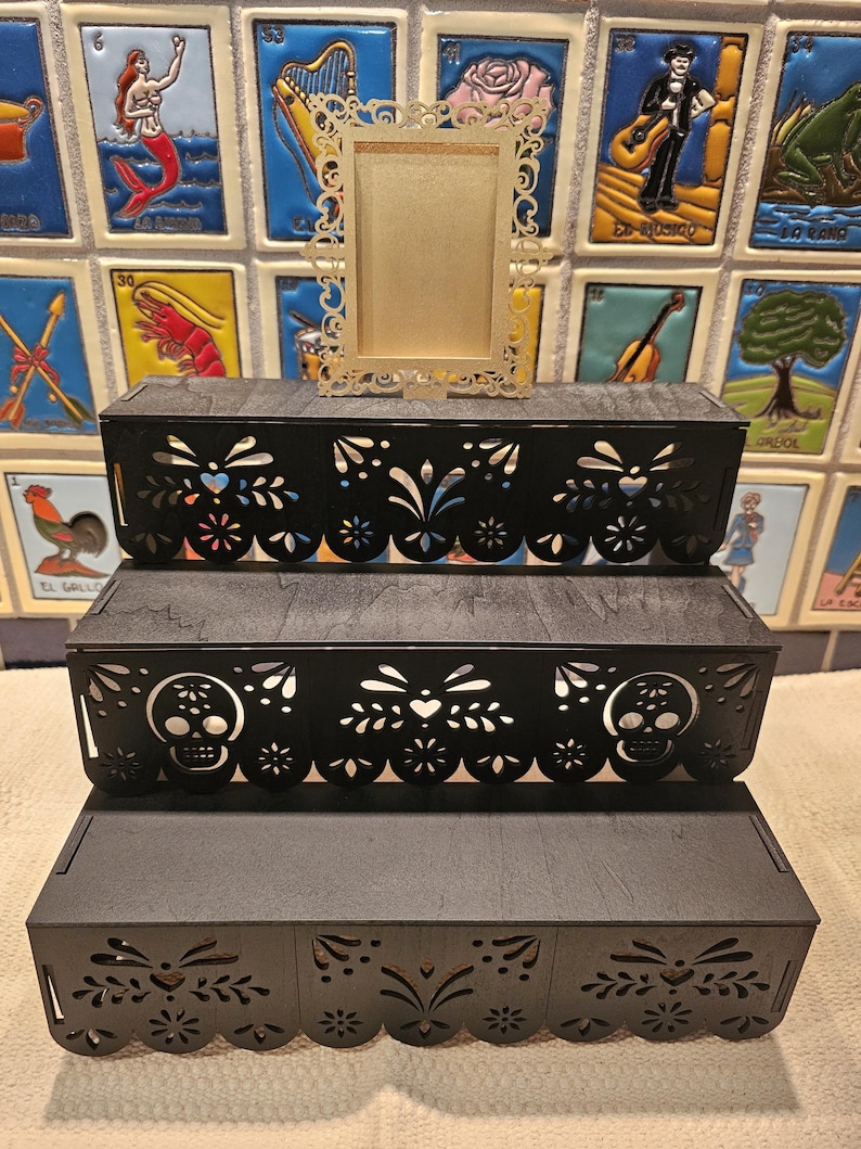 May include: A three-tiered black wooden display stand featuring intricate laser-cut patterns along the front edges, including skull motifs on the middle tier. A small, rectangular, gold-colored ornate frame sits centered on the top tier.