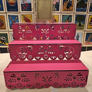 May include: A three-tiered pink display stand featuring intricate laser-cut patterns, including sugar skull motifs on the middle tier and scalloped edges. A small, ornate, rectangular gold-colored frame sits centered on the top tier of the stand.