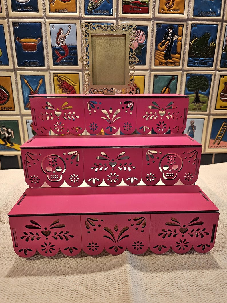 May include: A three-tiered pink display stand featuring intricate laser-cut patterns, including sugar skull motifs on the middle tier and scalloped edges. A small, ornate, rectangular gold-colored frame sits centered on the top tier of the stand.
