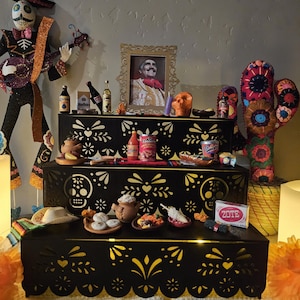 May include: A three-tiered black wooden display stand featuring intricate laser-cut patterns of skulls and floral motifs. The stand is decorated with miniature items including food cans, bottles, a framed portrait, a Zote soap bar, and a skeleton figure.