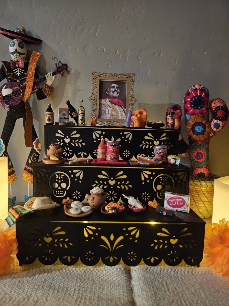 May include: A three-tiered black wooden display stand featuring intricate laser-cut patterns of skulls and floral motifs. The stand is decorated with miniature items including food cans, bottles, a framed portrait, a Zote soap bar, and a skeleton figure.