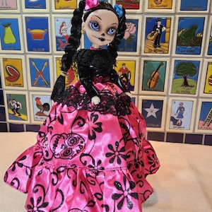 May include: A Day of the Dead doll with black braided hair, wearing a black lace top and a pink satin dress with black floral and skull patterns. The doll has blue eyes and is decorated with flowers.