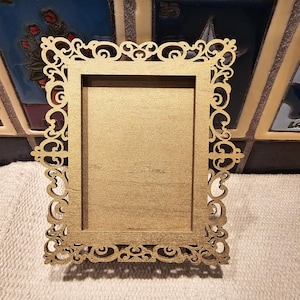 May include: A gold-colored, rectangular picture frame with an ornate, laser-cut design. The frame has a simple, rectangular opening for a photo and decorative scrollwork around the edges. The frame is made of wood.