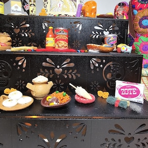 May include: A black tiered wooden display stand with intricate laser-cut floral and skull patterns. The shelves hold various miniature food items, a Zote soap bar, a framed portrait of a man, and small bottles, creating a detailed decorative arrangement.