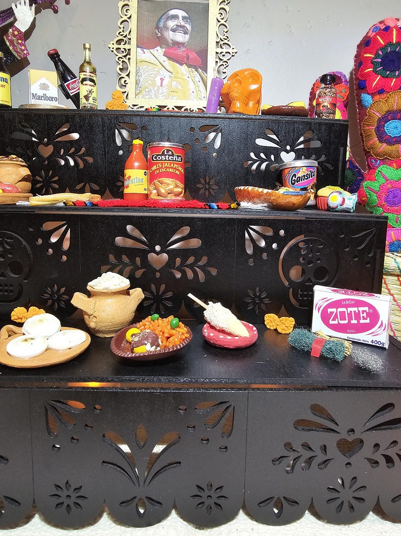 May include: A black tiered wooden display stand with intricate laser-cut floral and skull patterns. The shelves hold various miniature food items, a Zote soap bar, a framed portrait of a man, and small bottles, creating a detailed decorative arrangement.