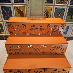 May include: A three-tiered orange wooden display stand featuring intricate laser-cut patterns, including skull motifs and floral designs along the front edges. A small, rectangular, ornate silver-colored frame sits centered on the top tier.