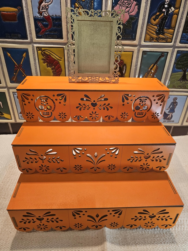 May include: A three-tiered orange wooden display stand featuring intricate laser-cut patterns, including skull motifs and floral designs along the front edges. A small, rectangular, ornate silver-colored frame sits centered on the top tier.