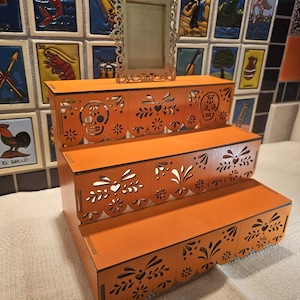 May include: A three-tiered orange wooden display stand featuring intricate laser-cut patterns of skulls and floral motifs along the front edges. A small, rectangular, ornate gold-colored frame stands upright on the top tier of the display unit.