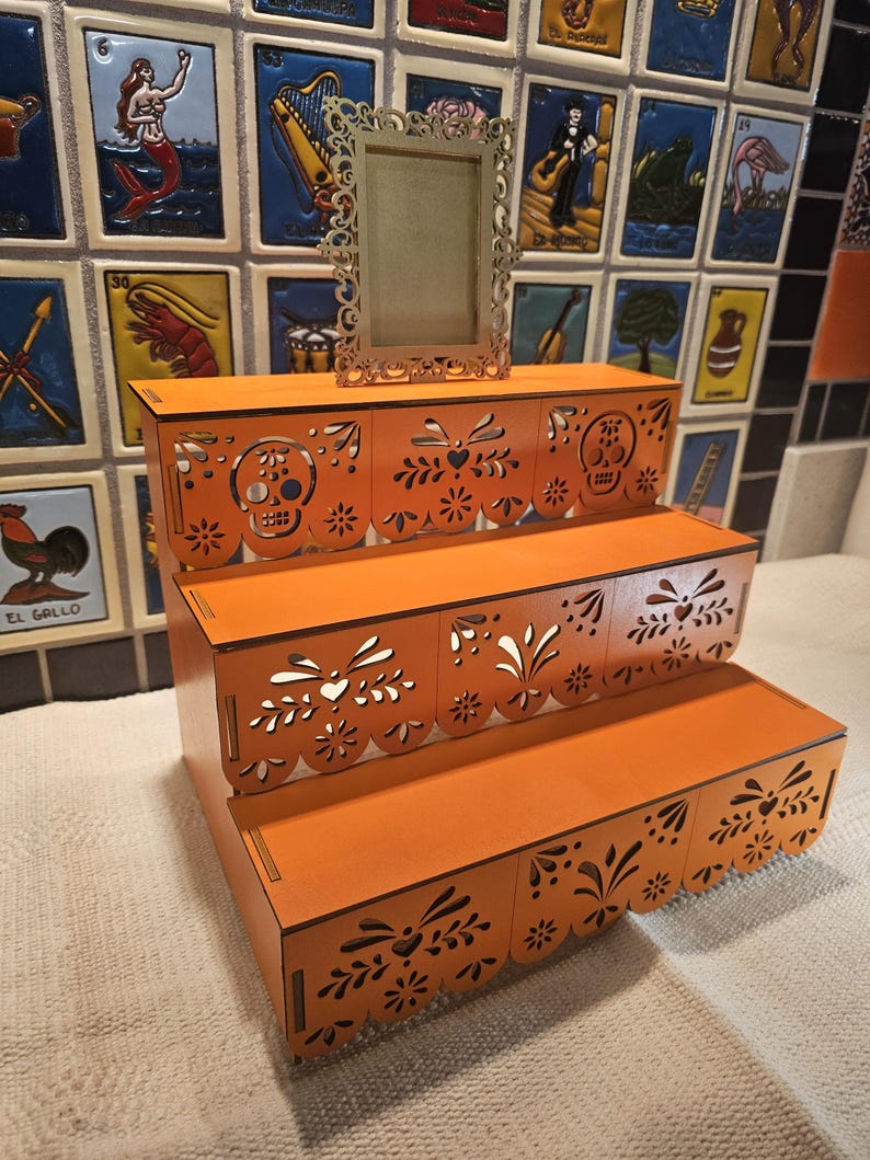 May include: A three-tiered orange wooden display stand featuring intricate laser-cut patterns of skulls and floral motifs along the front edges. A small, rectangular, ornate gold-colored frame stands upright on the top tier of the display unit.