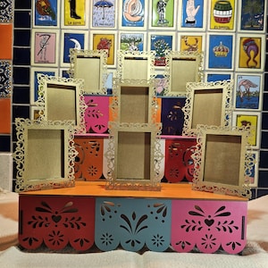 May include: A tiered display stand featuring a base with three colorful panels in red, blue, and pink, each with intricate laser-cut floral patterns. Nine gold-toned rectangular picture frames with ornate, filigree-style borders are arranged on top.