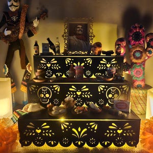 May include: A three-tiered black wooden display stand featuring intricate laser-cut sugar skull and floral patterns. The stand is illuminated from within and holds various miniature items, including food containers, bottles, and a framed portrait.