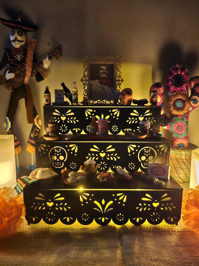 May include: A three-tiered black wooden display stand featuring intricate laser-cut sugar skull and floral patterns. The stand is illuminated from within and holds various miniature items, including food containers, bottles, and a framed portrait.