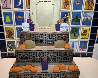 May include: A three-tiered wooden altar with decorative cutouts, featuring skull and candle accents. The altar is adorned with orange marigolds and a central wooden frame. The background showcases colorful Mexican Loteria tiles.