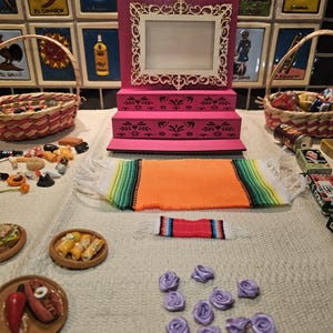 Peut inclure: Un stand rose à trois niveaux avec un cadre en bois décoratif, une couverture rayée colorée et des fleurs en tissu violet. Le stand est décoré de motifs floraux noirs.