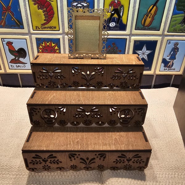 May include: A three-tiered wooden display stand with laser-cut decorative edges featuring floral patterns and skull motifs. A small, rectangular gold-colored frame with an ornate filigree border sits on the top tier of the display stand.