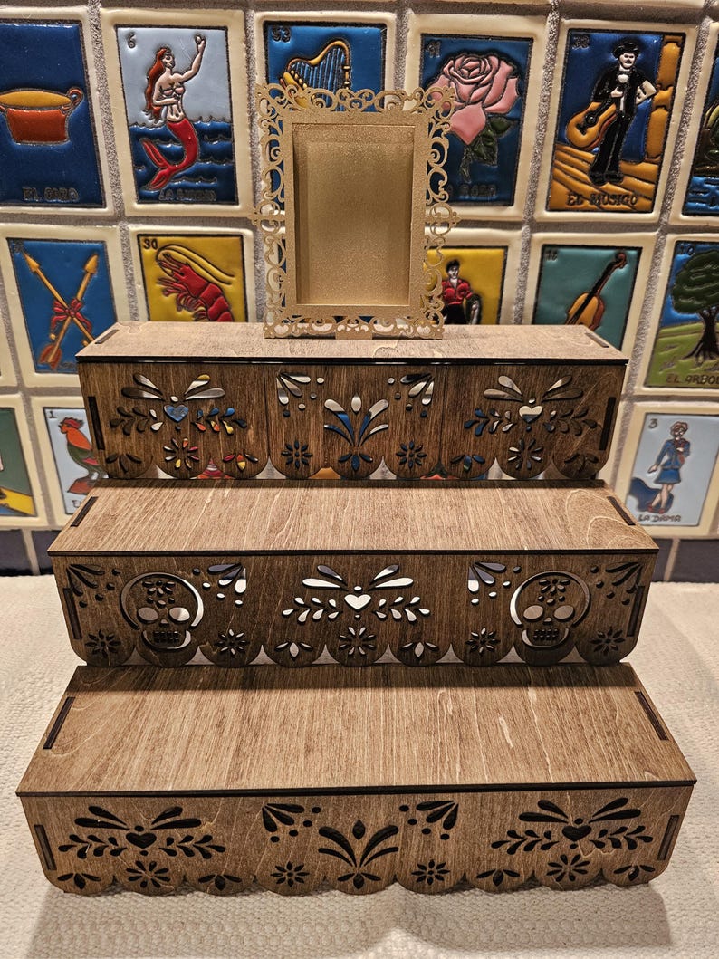 May include: A three-tiered wooden display stand with laser-cut decorative edges featuring floral patterns and skull motifs. A small, rectangular, gold-colored ornate frame with a filigree border sits centered on the top tier of the display.
