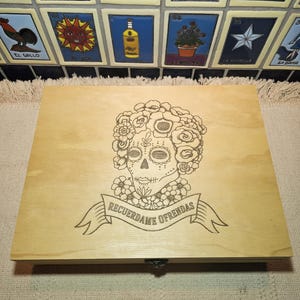 May include: A rectangular light-colored wooden box featuring a laser-engraved design of a sugar skull adorned with flowers. Below the skull, a banner contains the text "RECUERDAME OFRENDAS" in a clear, dark, capitalized font.