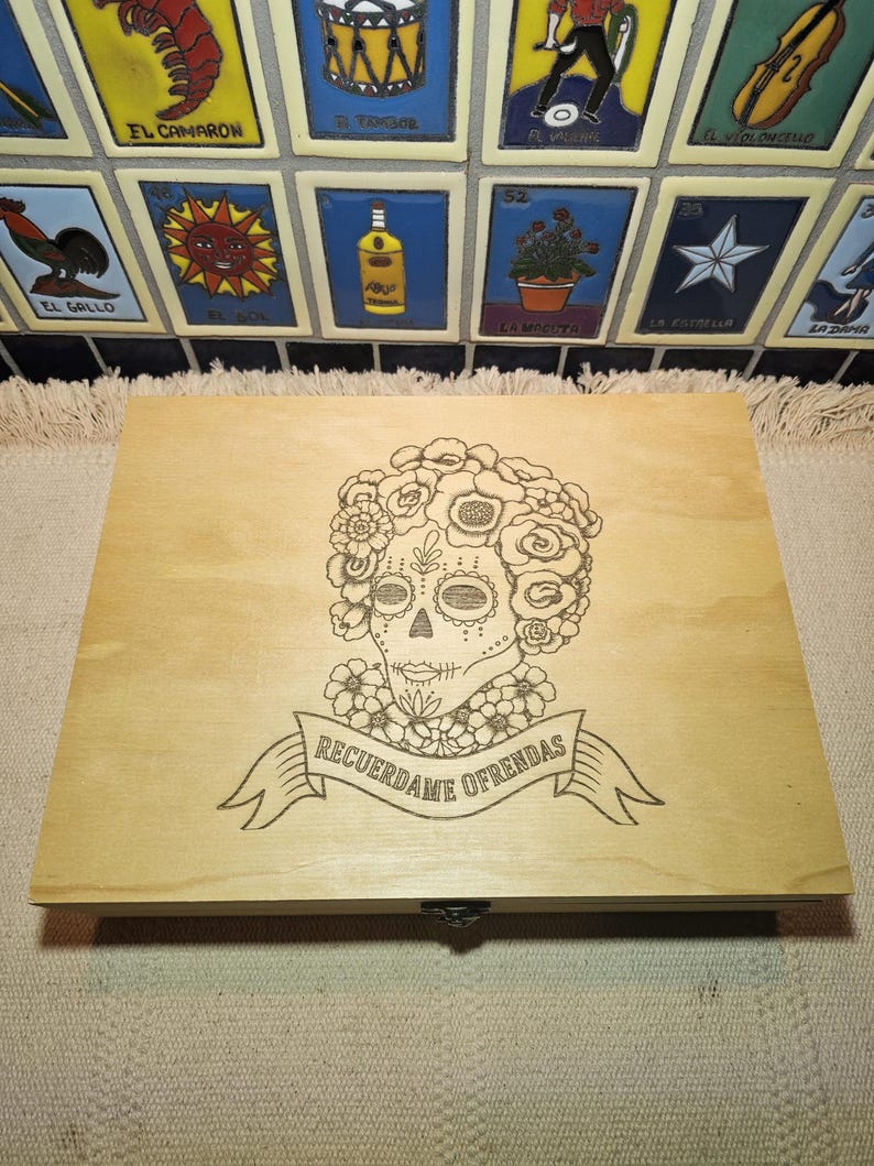 May include: A rectangular light-colored wooden box featuring a laser-engraved design of a sugar skull adorned with flowers. Below the skull, a banner contains the text "RECUERDAME OFRENDAS" in a clear, dark, capitalized font.