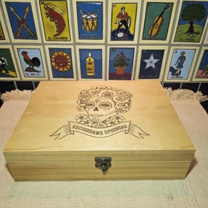 May include: A rectangular light-colored wooden box with a hinged lid and a metal latch. The lid features a laser-engraved design of a sugar skull surrounded by flowers, with a banner below containing the text RECUERDAME OFRENDAS.