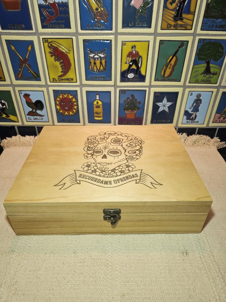 May include: A rectangular light-colored wooden box with a hinged lid and a metal latch. The lid features a laser-engraved design of a sugar skull surrounded by flowers, with a banner below containing the text RECUERDAME OFRENDAS.