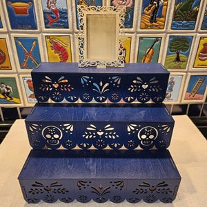 May include: A three-tiered dark blue wooden display stand featuring intricate laser-cut patterns along the front edges, including skull motifs on the middle tier. A small, rectangular, light-colored wooden frame with ornate filigree edges sits on top.