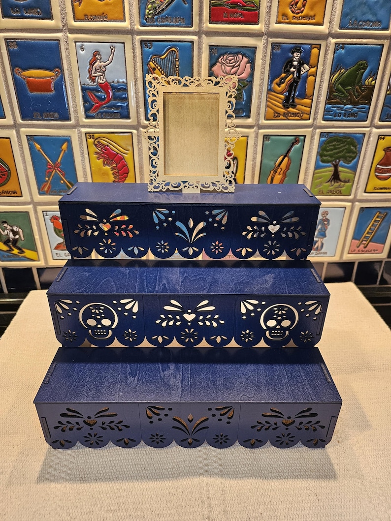 May include: A three-tiered dark blue wooden display stand featuring intricate laser-cut patterns along the front edges, including skull motifs on the middle tier. A small, rectangular, light-colored wooden frame with ornate filigree edges sits on top.