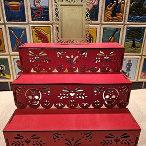 May include: A three-tiered red wooden display stand featuring intricate laser-cut patterns, including skull motifs on the middle tier and scalloped edges. A small, rectangular, gold-colored frame with an ornate, filigree-style border sits on top.