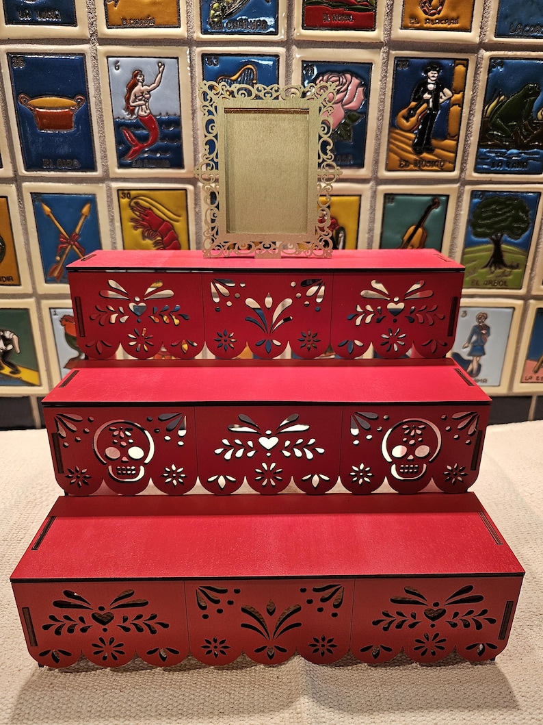 May include: A three-tiered red wooden display stand featuring intricate laser-cut patterns, including skull motifs on the middle tier and scalloped edges. A small, rectangular, gold-colored frame with an ornate, filigree-style border sits on top.