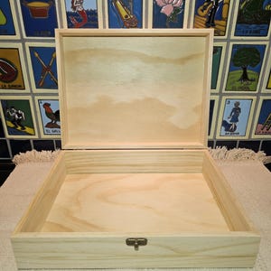 May include: A square, light-colored wooden box with an open lid, featuring a natural wood grain finish and a small, centered metal latch on the front edge. The box is constructed from smooth, unfinished wood with visible hinges at the back.