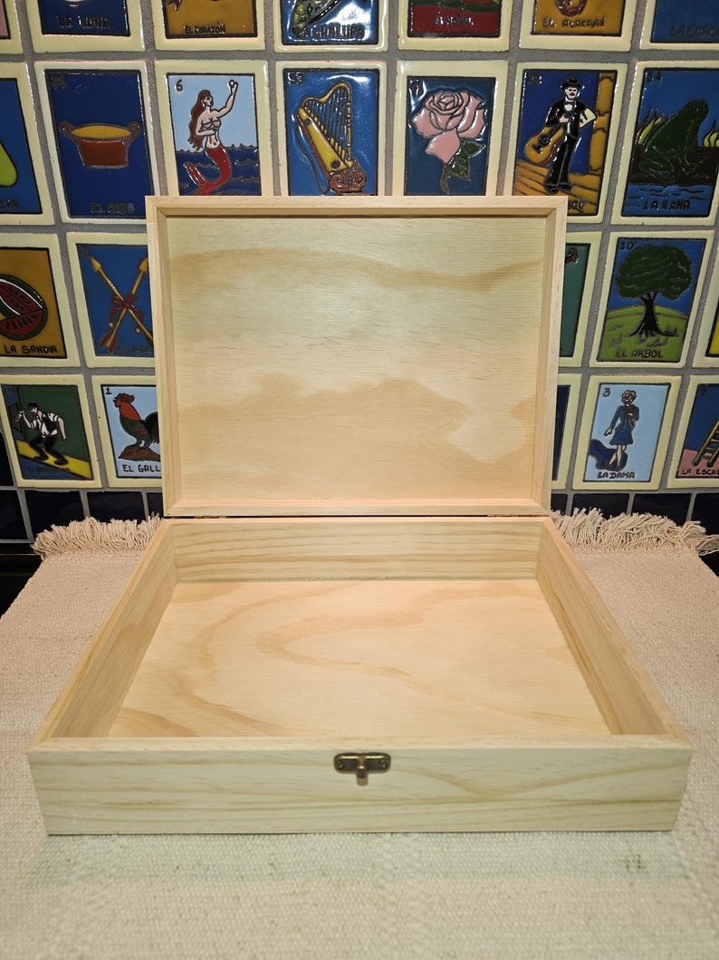 May include: A square, light-colored wooden box with an open lid, featuring a natural wood grain finish and a small, centered metal latch on the front edge. The box is constructed from smooth, unfinished wood with visible hinges at the back.