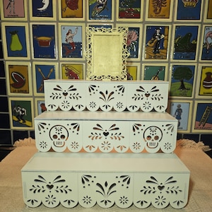 May include: A three-tiered white wooden display stand featuring intricate laser-cut patterns resembling papel picado, including skull motifs on the middle tier. A small, ornate, rectangular white frame sits centered on the top tier of the stand.