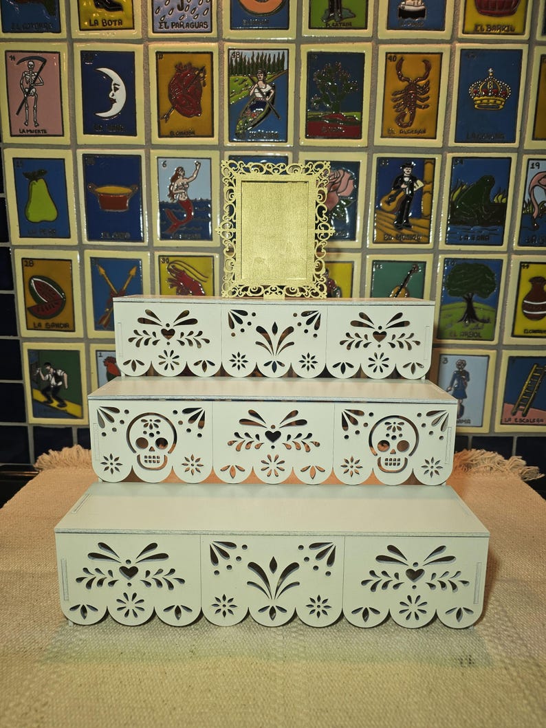 May include: A three-tiered white wooden display stand featuring intricate laser-cut patterns resembling papel picado, including skull motifs on the middle tier. A small, ornate, rectangular white frame sits centered on the top tier of the stand.