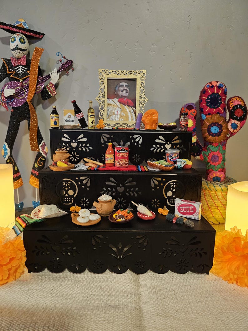 May include: A three-tiered black wooden display stand with intricate cut-out patterns, featuring a collection of miniature food items, a framed portrait of a man in traditional attire, a skeleton figure, a fabric cactus, and Zote soap.