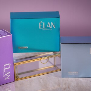 Könnte beinhalten: Drei Schachteln mit Hautpflegeprodukten von Elan Professional Lab. Die Schachteln sind übereinander gestapelt. Die oberste Schachtel ist türkisblau, die mittlere Schachtel ist aus klarem Glas mit goldener Einfassung und die unterste Schachtel ist hellblau.