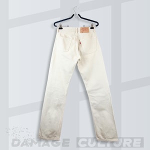 May include: Off-white denim jeans hanging on a black plastic hanger. The jeans feature a tan leather patch on the back waistband, two back pockets, and a small red tab on the right pocket. The design includes a button closure and belt loops.