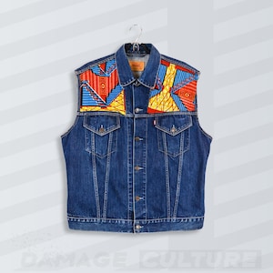 May include: A blue denim vest with colourful patterned fabric on the shoulders. The vest has a classic collar, two chest pockets, and a button-up front. The patterned fabric features red, yellow, and blue geometric shapes.