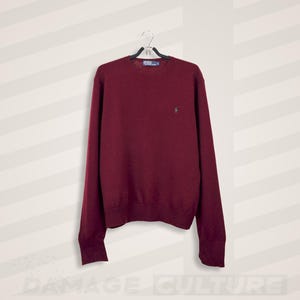 May include: A burgundy crew neck jumper hanging on a black hanger. The jumper has long sleeves and a small logo on the chest. The background is a white wall with diagonal stripes.