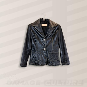 Peut inclure: Veste en cuir noir avec un col cranté, deux poches avant et deux boutons argentés. La veste a des coutures dorées et est suspendue à un cintre en métal.