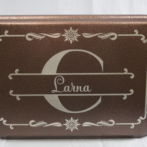 May include: A brown metal cake pan with a silver engraved design. The design features a large letter "C" with the name "Lorna" written below it. The pan has a decorative border with a floral design.