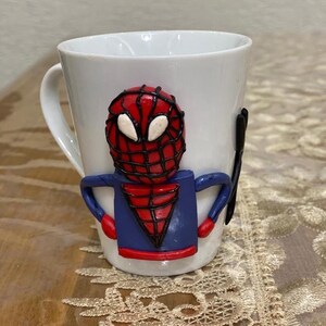 以下が含まれることがあります： 白いセラミック製のマグカップで、前面に赤と青の3D粘土製のスパイダーマンのフィギュアが付いています。フィギュアは黒いウェブデザインになっています。